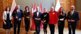 W sali z wyłożonymi drewnianymi panelami ścianami stoi w szeregu grupa ośmiu osób, w rękach trzymają po jednej czerwonej róży, za nimi pod ścianą polskie flagi.