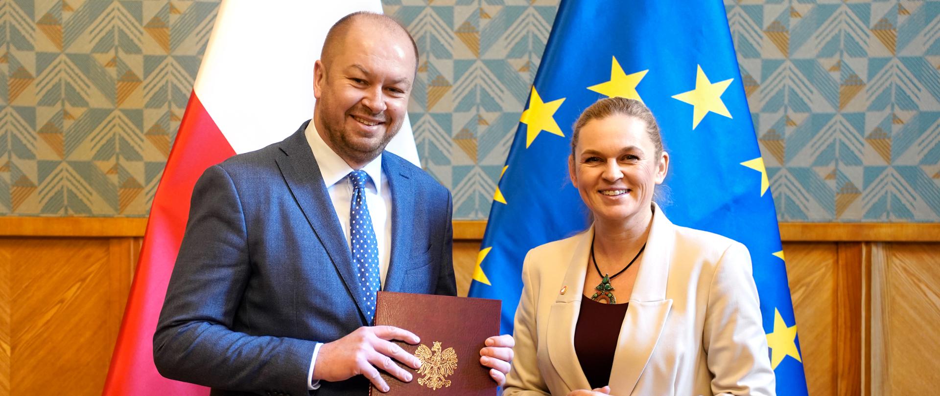 Mężczyzna w garniturze stoi obok minister Nowackiej w ręku trzyma teczkę z godłem państwowym. W tle flaga biało czerwona i flaga unijna.