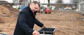 Zdjęcie na dworze, minister Czarnek stoi przy stoliku na którym leży wydrążony biały czworokątny kamień, w ręku trzyma kielnię, za nim ogrodzenie z siatki i pryzma piasku.