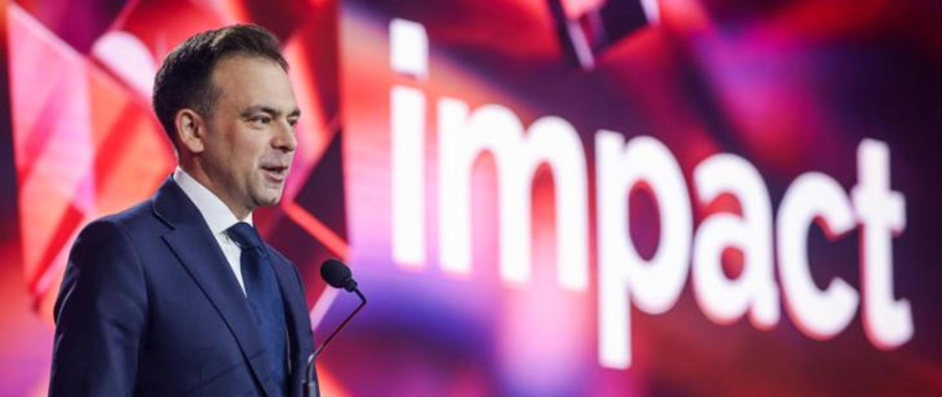  Minister finansów i gospodarki Andrzej Domański przy mikrofonie na scenie podczas wydarzenia „Impact”, na tle dużego napisu impact i czerwono‑różowej scenografii świetlnej.