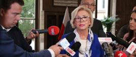 Wiceminister Machałek w niebieskim ubraniu stoi i mówi, dookoła niej dziennikarze trzymający mikrofony.