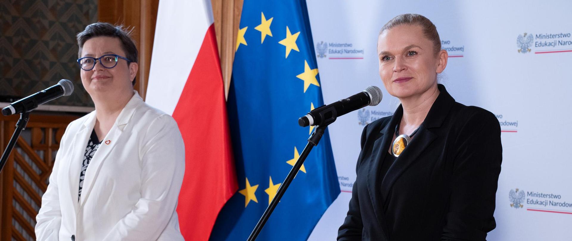 Widok z boku. Na pierwszym planie minister Barbara Nowacka stoi przy mikrofonie i słucha, obok niej wiceminister Katarzyna Lubnauer. W tle ścianka z napisem Ministerstwo Edukacji Narodowej oraz flagi Polski i UE.