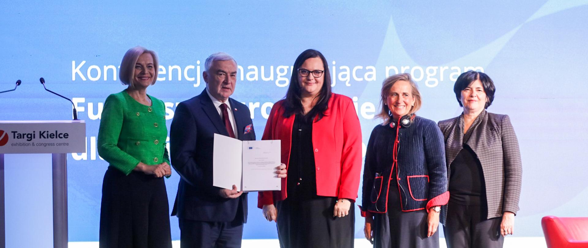 Na zdjęciu widocznych jest 5 osób (4 kobiety i mężczyzna). W środku stoi Sekretarz Stanu Małgorzata Jarosińska-Jedynak, w tle ekran konferencji.