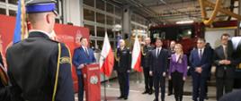 W hali garażowej stoi grupa kilku osób, minister Czarnek stoi przy mównicy, przed nim mężczyźni w czarnych galowych mundurach, w tle samochód pożarniczy.