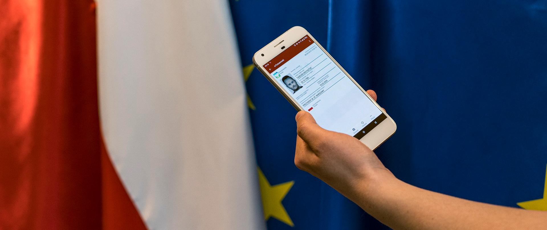 W tle flagi. Na pierwszym planie ręka ze smartfonem. Na ekranie aplikacja mObywatel.
