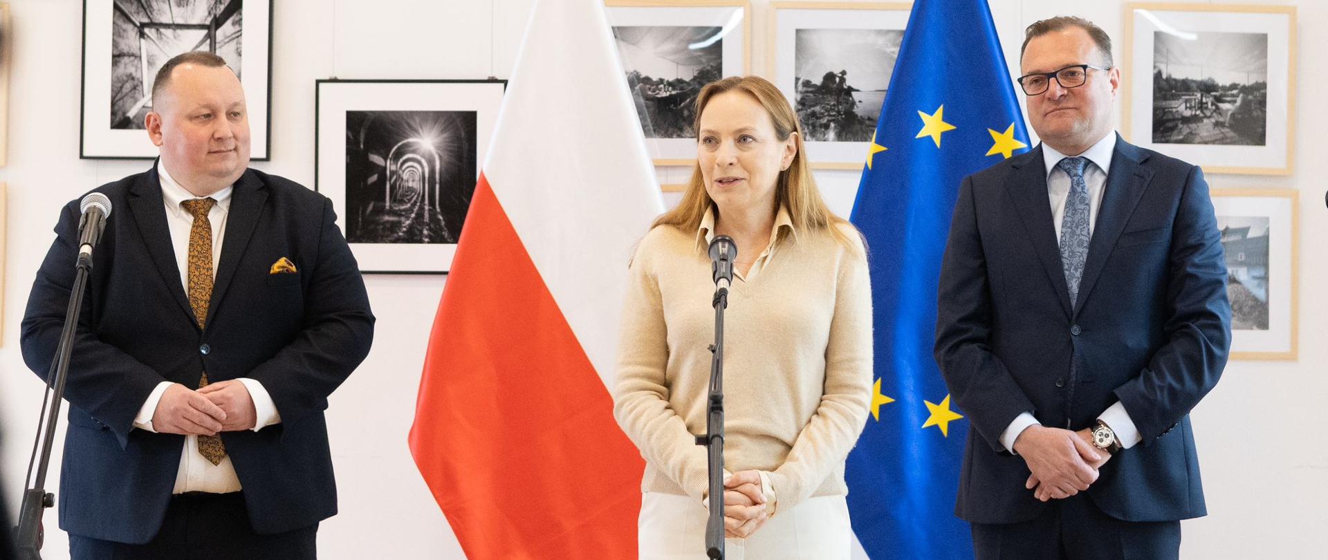 Trzy osoby stoją obok siebie. W środku przy mikrofonie minister Katarzyna Pełczyńska-Nałęcz. W tle za ich plecami flagi Polski i Unii Europejskiej. 