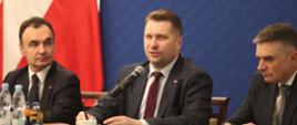 Przy nakrytym ciemnoszarym materiałem stole siedzi minister Czarnek i dwóch mężczyzn w garniturach, minister mówi do mikrofonu, za nimi niebieska ściana, z lewej polska flaga.