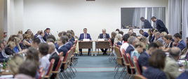 Widok na salę, na której przy dwóch długich stołach ustawionych wzdłuż sali siedzą osoby. Na końcu sali ustawiony jest trzeci stół, który znajduję się prostopadle do dwóch pozostałych, siedzi przy nim minister Kwieciński. 