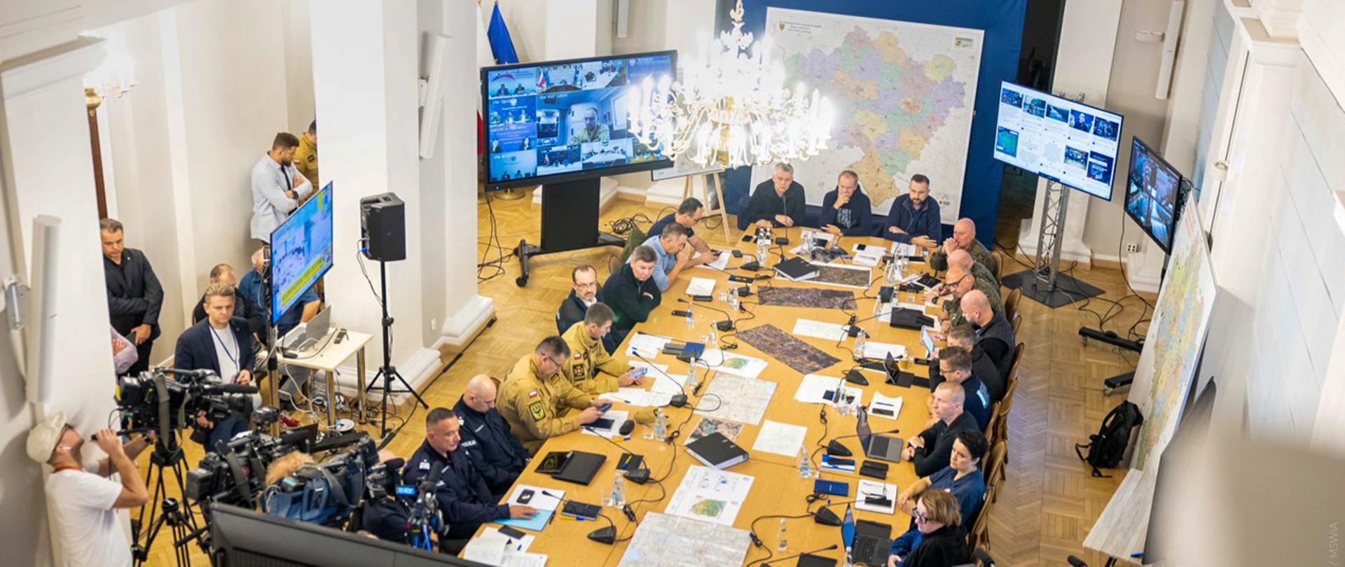 Na zdjęciu zrobionym z góry siedzą uczestnicy narady przy dużym stole na którym lezą mapy i sprzęt elektroniczny 