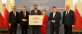 Pod szarą ścianą z napisem Śląski Urząd Wojewódzki w Katowicach stoi pięciu mężczyzn w garniturach i kobieta w czerwonym ubraniu, mężczyzna pośrodku grupy trzyma białą tabliczkę z napisem Branżowe Centrum Umiejętności.
