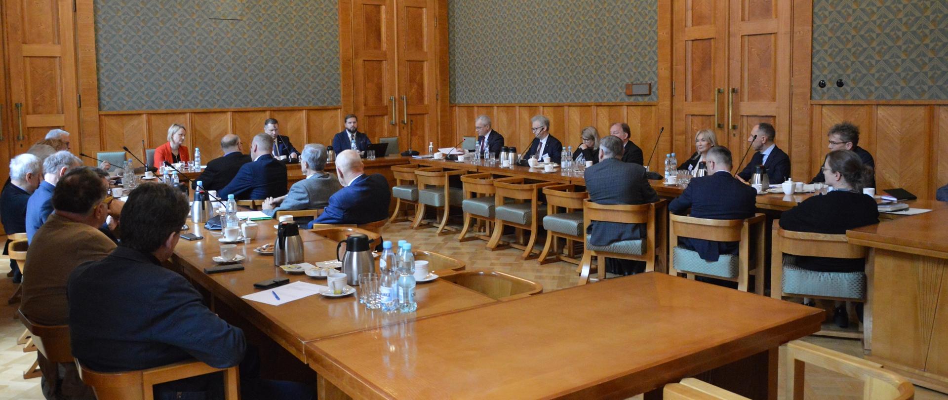 Widok z boku na salę z zielonymi ścianami zdobionymi drewnianymi panelami, przy ustawionych w czworokąt stołach siedzą ludzie, pod sufitem kryształowy żyrandol.