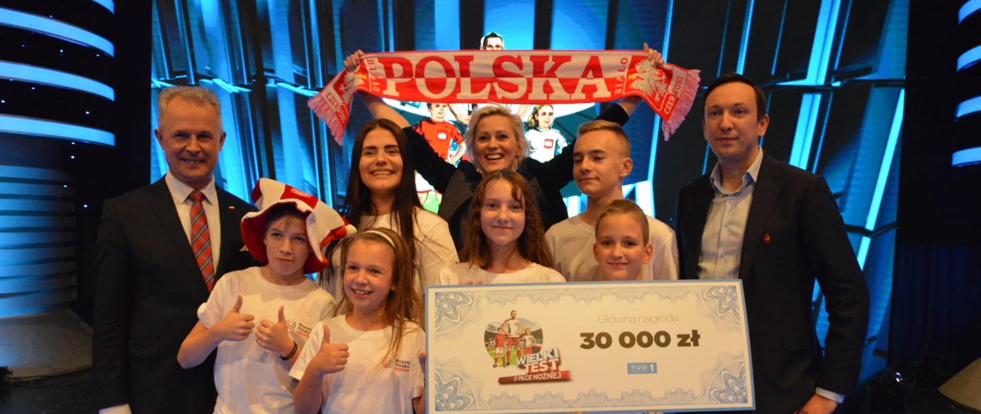 Oświetlone na niebiesko studio, na lustrzanej podłodze stoi grupa ludzi, w środku kilkoro dzieci w białych koszulkach trzyma tabliczkę z napisem 30 000 zł, za nimi kobieta trzyma rozpostarty biało-czerwony szalik z napisem Polska, po bokach dwóch mężczyzn w garniturach.