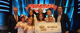 Oświetlone na niebiesko studio, na lustrzanej podłodze stoi grupa ludzi, w środku kilkoro dzieci w białych koszulkach trzyma tabliczkę z napisem 30 000 zł, za nimi kobieta trzyma rozpostarty biało-czerwony szalik z napisem Polska, po bokach dwóch mężczyzn w garniturach.
