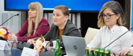 Na zdjęciu widać siedzące przy stole prezydialnym (od lewej): Rzecznik Praw Dziecka Monikę Horną-Cieślak, wiceminister w MEN Paulinę Piechnę-Więckiewicz oraz wicemarszałek Senatu Magdalenę Biejat. W trakcie wykonywania zdjęcia wiceminister Paulina Piechna-Więckiewicz przemawia do włączonego mikrofonu. Na stole widać również szklanki, butelki z wodą, identyfikatory imienne, pozostałe mikrofony oraz bukiety kwiatów. W tle widać ekran z niebieskim tłem.