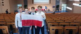 Widok od tyłu na salę, w przejściu między rzędami pustych krzeseł stoi czterech młodych mężczyzn którzy razem trzymają polską flagę.