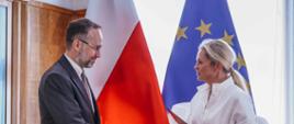 Mężczyzna i kobieta uściskiem dłoni przypieczętowują oficjalne spotkanie. Mężczyzna, w okularach i garniturze, z brodą, uśmiecha się, patrząc na kobietę. Kobieta, z włosami spiętymi w kok, ubrana jest w białą, luźną sukienkę z szerokimi rękawami. W lewej ręce trzyma czerwoną teczkę, a na lewym nadgarstku ma sportowy zegarek. W tle widoczne są flagi Polski i Unii Europejskiej. Atmosfera jest formalna i uroczysta.