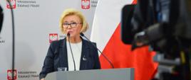 Wiceminister stoi przy mównicy z orłem, w tle biało-czerwona flaga. Na pierwszym planie kamery 
