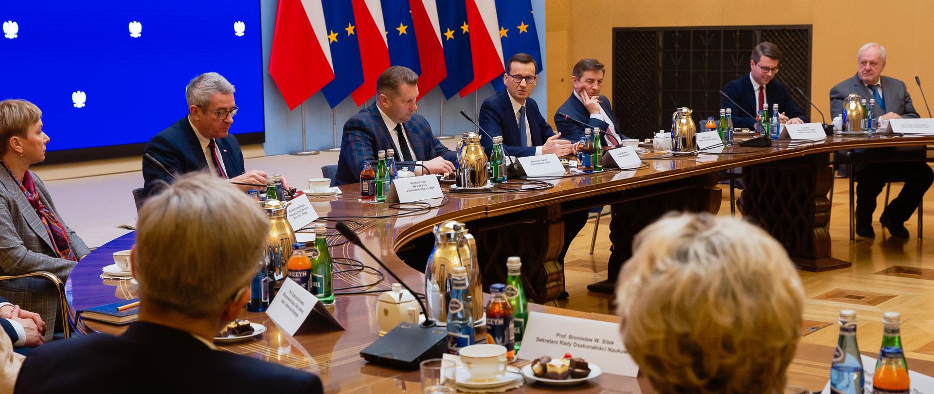 Widok z boku na sale, w której przy ustawionym w prostokąt stole siedzą ludzie w garniturach. Za nimi pod ścianą stoją flagi Polski i UE.