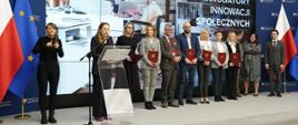 Minister funduszy i polityki regionalnej Katarzyna Pełczyńska-Nałęcz i wiceministra Monika Sikora z beneficjenci konkursu na inkubatory na innowacje społeczne, uczestnicy stoją na tle zdjęcia przedstawiającego młodego mężczyznę na wózku inwalidzkim