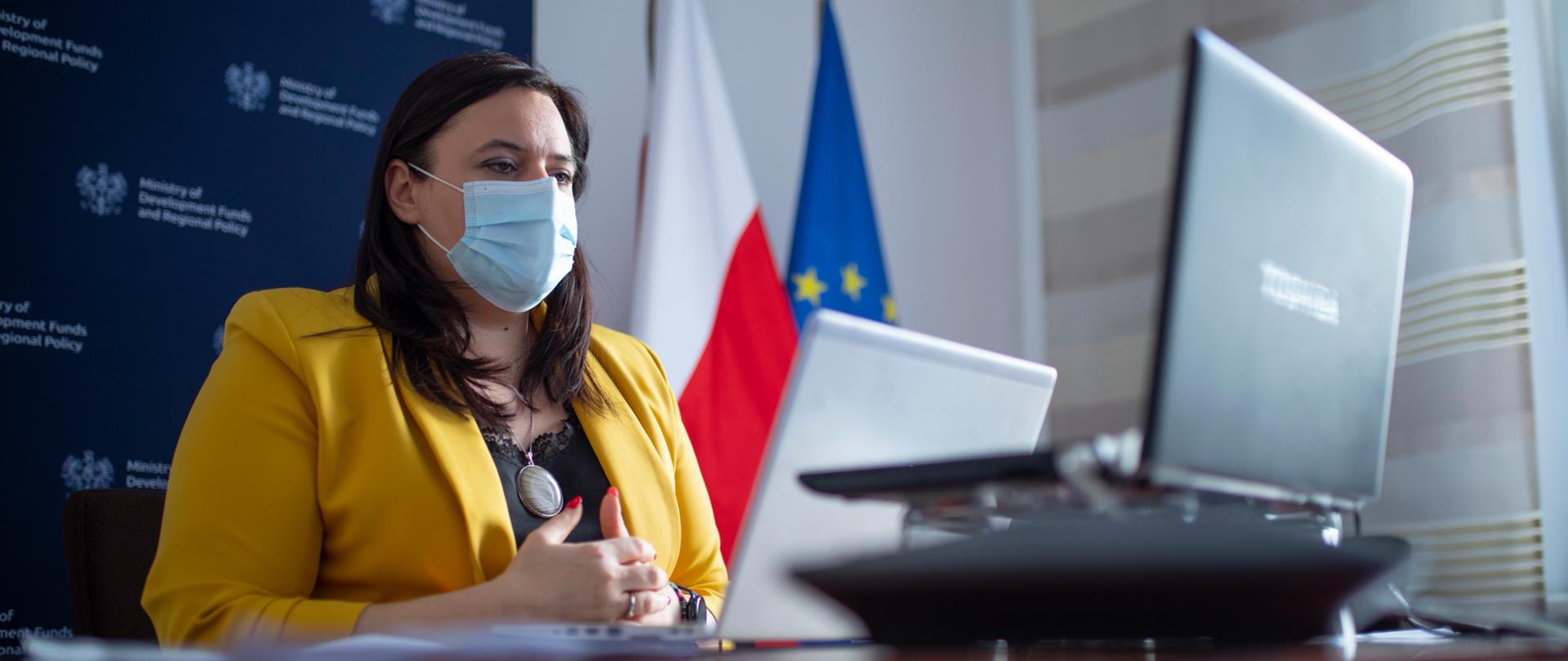 Wiceminister Małgorzata Jarosińska-Jedynak za maseczką na twarzy siedzi przy biurku przed laptopem. Za nią ścianka promocyjna Ministerstwa Funduszy i Polityki Regionalnej oraz dwie flagi PL i UE.