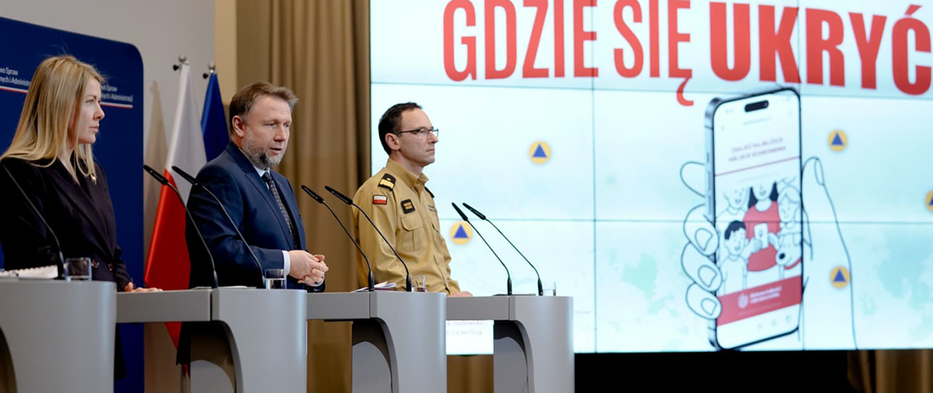 Kierownictwo MSWiA i komendant główny PSP przy mównicach. W tle telebim z napisem "Gdzie się ukryć" i zdjęciem telefonu z aplikacją nt. schronów
