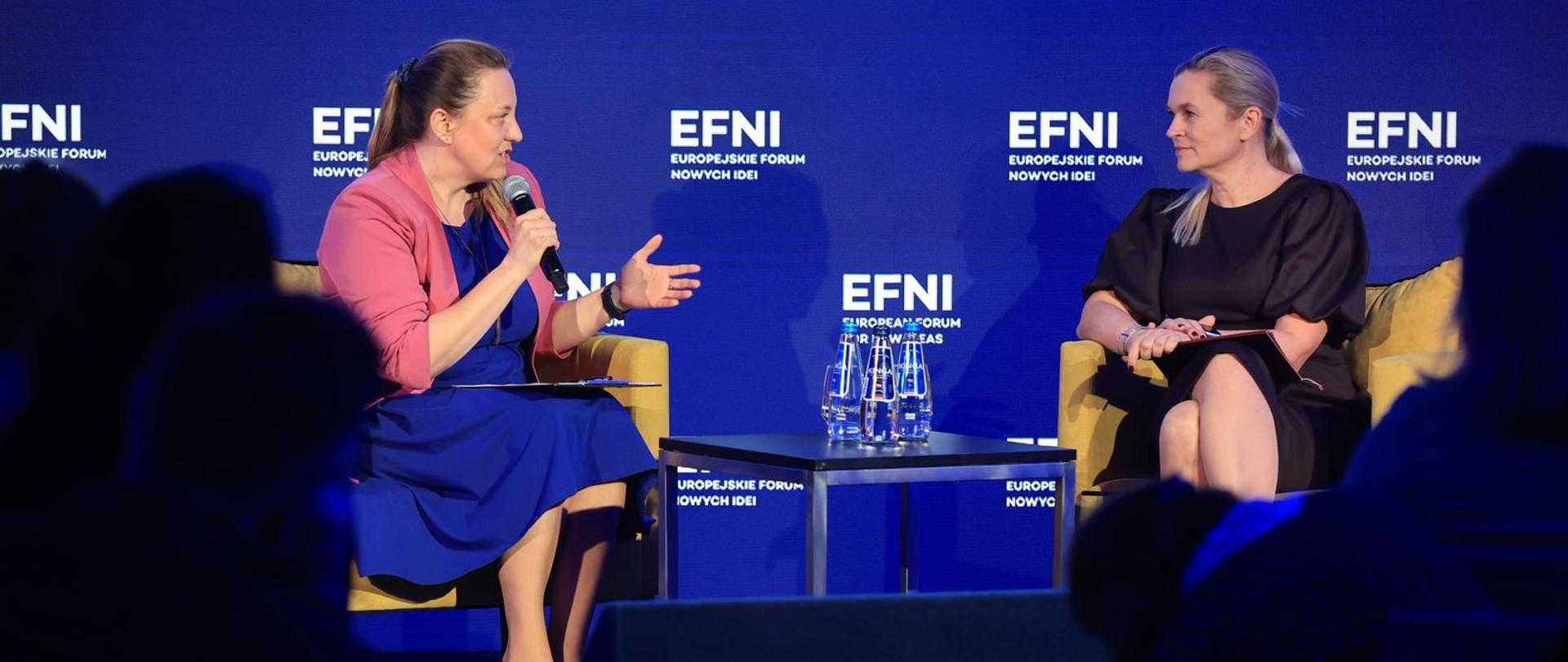 Na scenie siedzą dwie kobiety, pomiędzy nimi stoi stolik na którym ustawiono butelki z wodą. W tle niebieska ścianka z napisem EFNI Europejskie Forum Nowych Idei 