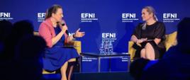 Na scenie siedzą dwie kobiety, pomiędzy nimi stoi stolik na którym ustawiono butelki z wodą. W tle niebieska ścianka z napisem EFNI Europejskie Forum Nowych Idei 