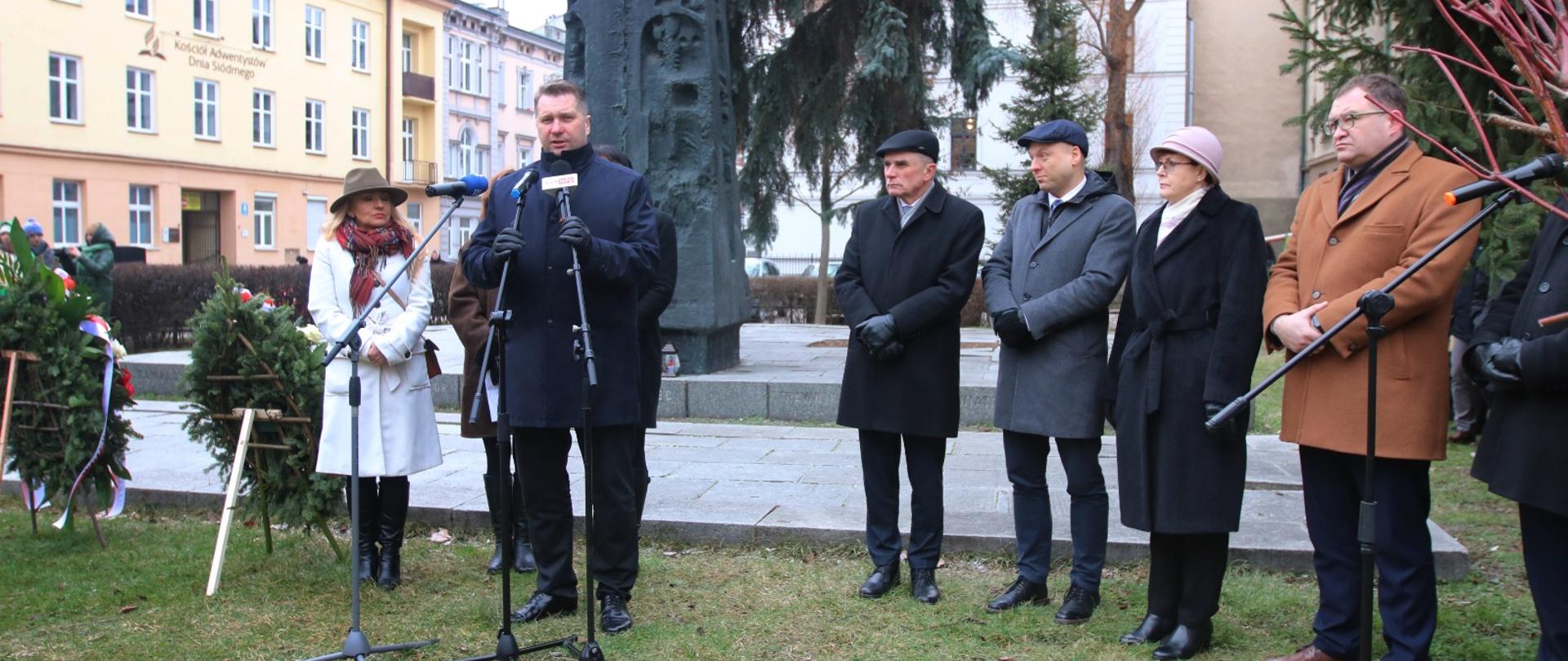 Minister Czarnek w otoczeniu kilku osób stoi na dworze na trawniku i mówi do kilku mikrofonów na stojakach, za nim czarny pomnik na środku placu.