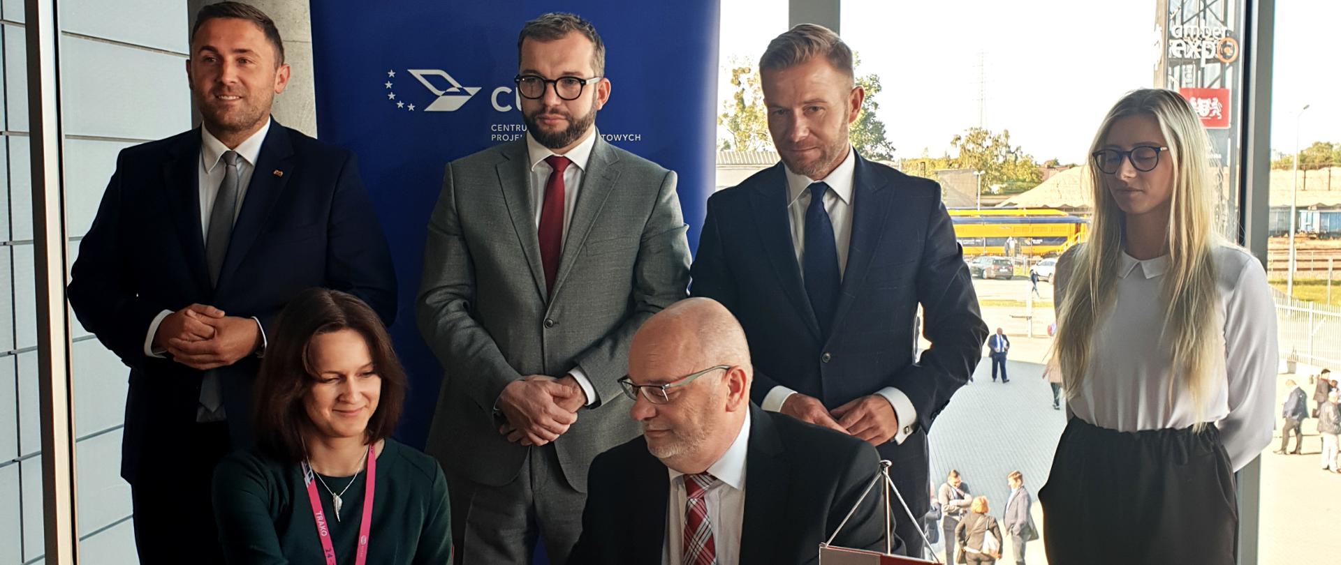 Minister Puda stoi przy stole, przy którym podpisywana jest umowa, wokół ministra stoją pozostali uczestnicy spotkania 