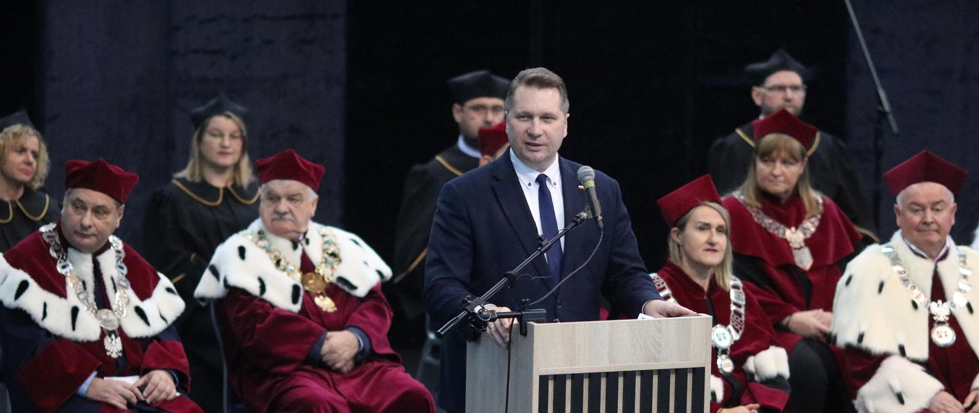 Minister Czarnek stoi przy mównicy i mówi do mikrofonu, za nim siedzą ludzie w ceremonialnych czerwonych strojach.