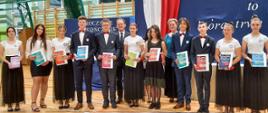 Na sali gimnastycznej stoi grupa kilkunastu elegancko ubranych osób, trzymają książki, za nimi wiceminister Bernacki.
