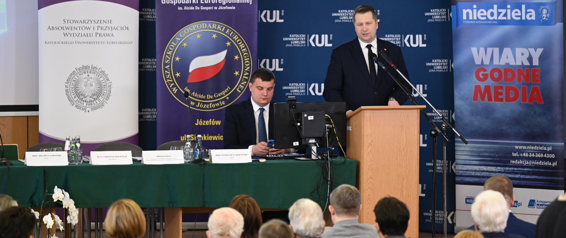 minister czarnek przemawia na rozpoczęcie konferencji. Ubrany w granatową marynarkę, białą koszulę i granatowy krawat. w tle ścianka uniwersytetu z napisem KUL. zdjęcie przedstwia szerszy kadr, przed przemawiającym ministrem widać zgromadzonych uczestników konferencji