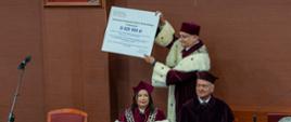 Za nakrytym czerwonym materiałem stołem siedzi kobieta w czerwonym ceremonialnym stroju i mężczyzna w czarnym ubraniu, za nimi stoi mężczyzna w czerwonym stroju trzymający w rękach wielki symboliczny czek z napisem 31 825 500 zł.