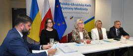 Przy białym stole siedzi kilka osób, za nimi flagi Polski, Ukrainy i UE, za nimi na ściance napis Polsko-Ukraińska Rada Wymiany Młodzieży.