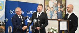 Minister Czarnek stoi pod ścianą na której wisi rodzinny portret, obok niego dwóch mężczyzn w garniturach, jednemu minister podaje rękę, drugi trzyma oprawiony w ramki dokument, przed nimi kilka mikrofonów.