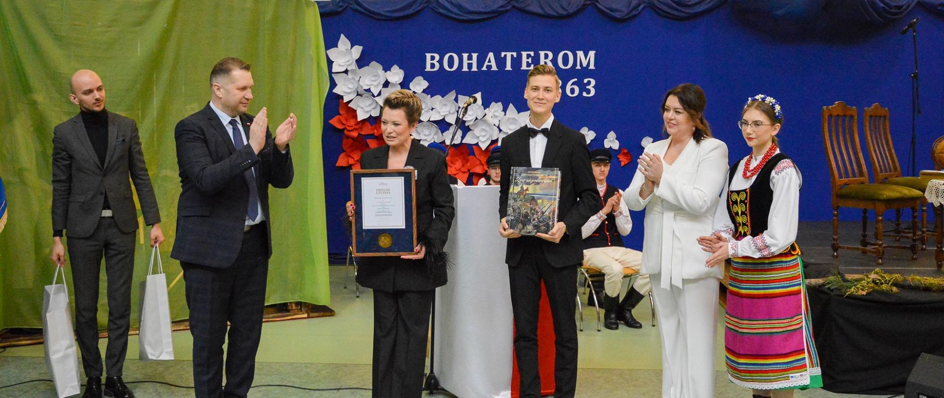 Na tle częściowo przysłoniętej zielonym materiałem niebieskiej ściany z napisem Bohaterom 1863 stoi sześć osób, wśród nich minister Czarnek który bije brawo, obok niego ubrana na czarno kobieta i młody człowiek w czarnym garniturze, oboje trzymają oprawione w ramki dokumenty.