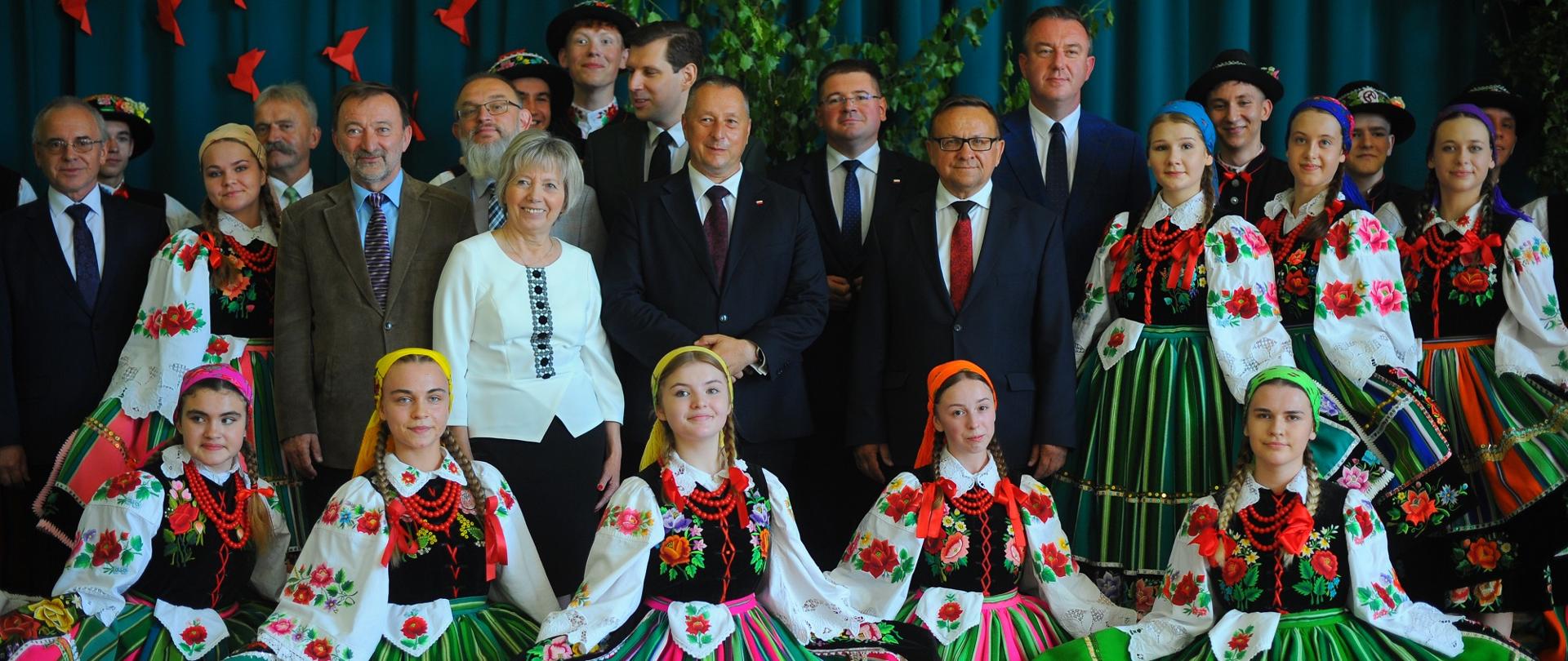 Uczestnicy wizyty i grupa uczennic w ludowych strojach stoją pod ścianą sali gimnastycznej.