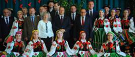 Uczestnicy wizyty i grupa uczennic w ludowych strojach stoją pod ścianą sali gimnastycznej.