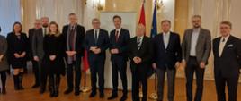 W eleganckiej sali z żółtymi ścianami i drewnianą błyszczącą podłogą stoi duża grupa elegancko ubranych ludzi.