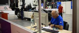 Mężczyzna w niebieskiej koszulce siedzi przy laptopie leżącym na stoliku, w tle wnętrze dużej hali w której stoi ciągnik siodłowy.