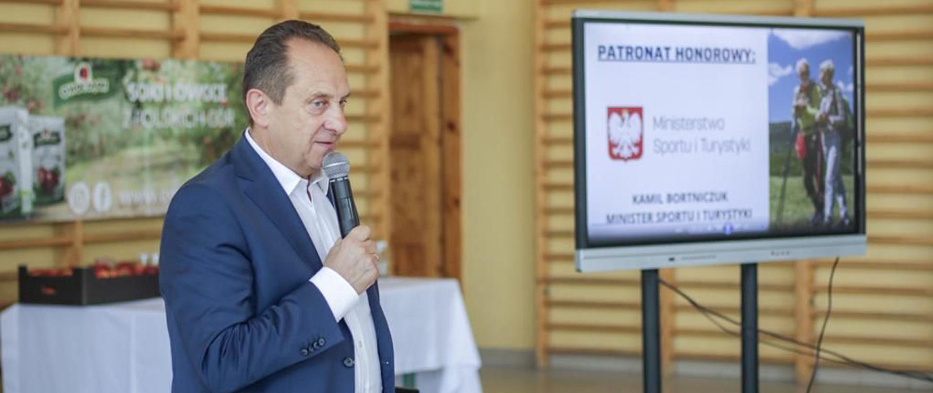 Na zdjęciu znajduje się elegancko ubrany mężczyzna w średnim wieku. Mężczyzna ten jest w trakcie wystąpienia publicznego, trzymając w prawym ręku mikrofon. W tle, po prawej stronie zdjęcia znajduje się ekran multimedialny, na którym wyświetlona jest plansza z logotypem ministerstwa sportu i turystyki oraz napisy "patronat honorowy" i "Kamil Bortniczuk, Minister Sportu i Turystyki". Zdjęcie wykonano od pasa w górę. 