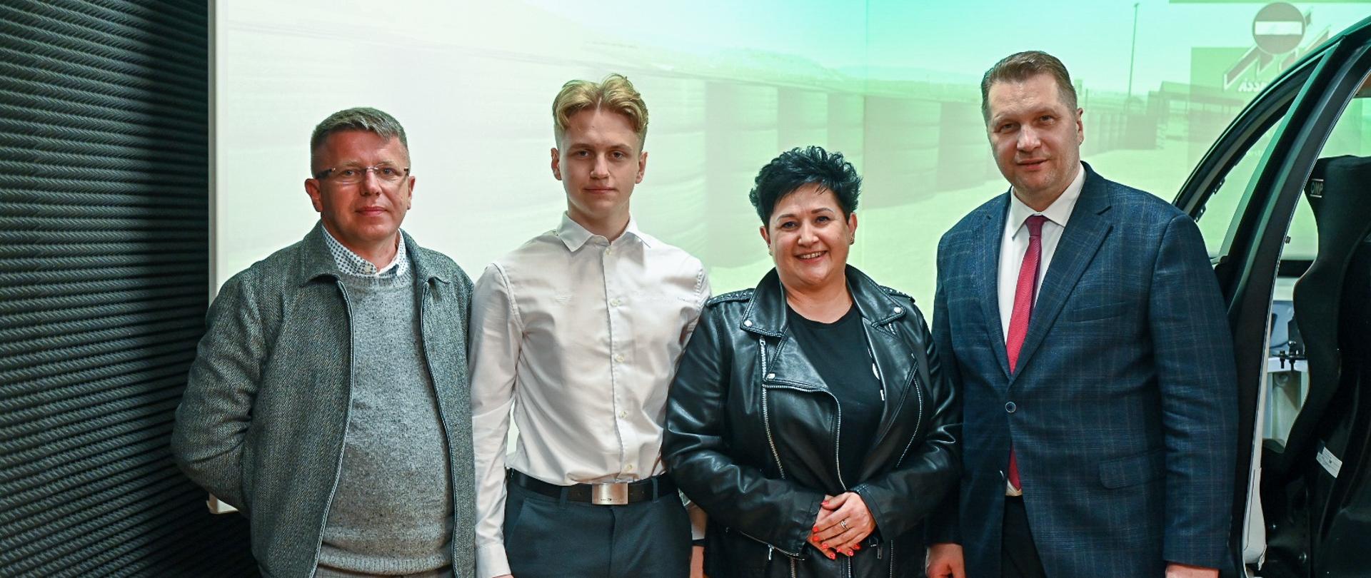 Minister Czarnek, młody mężczyzna w białej koszuli, kobieta w czarnym ubraniu i mężczyzna w szarym garniturze stoją w rzędzie, obok nich połowa przeciętego w poprzek samochodu.
