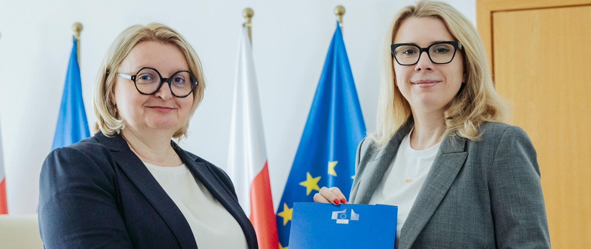 Wiceministra funduszy i polityki regionalnej Monika Sikora trzymają z dyrektor Katariną Ivanković Knežević z Dyrekcji Generalnej ds. Zatrudnienia, Spraw Społecznych i Włączenia Społecznego Komisji Europejskiej niebieską teczkę, za ich plecami flagi Polski i UE