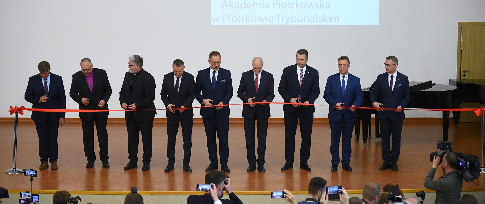 Na drewnianej podłodze dużej sali stoi w szeregu grupa mężczyzn w garniturach, każdy trzyma nożyczki i tnie kawałek przedzielającej salę czerwonej wstążki, za nimi jasnoszara ściana na której wisi godło, obok którego wyświetlany jest z rzutnika napis AP.