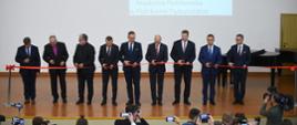 Na drewnianej podłodze dużej sali stoi w szeregu grupa mężczyzn w garniturach, każdy trzyma nożyczki i tnie kawałek przedzielającej salę czerwonej wstążki, za nimi jasnoszara ściana na której wisi godło, obok którego wyświetlany jest z rzutnika napis AP.