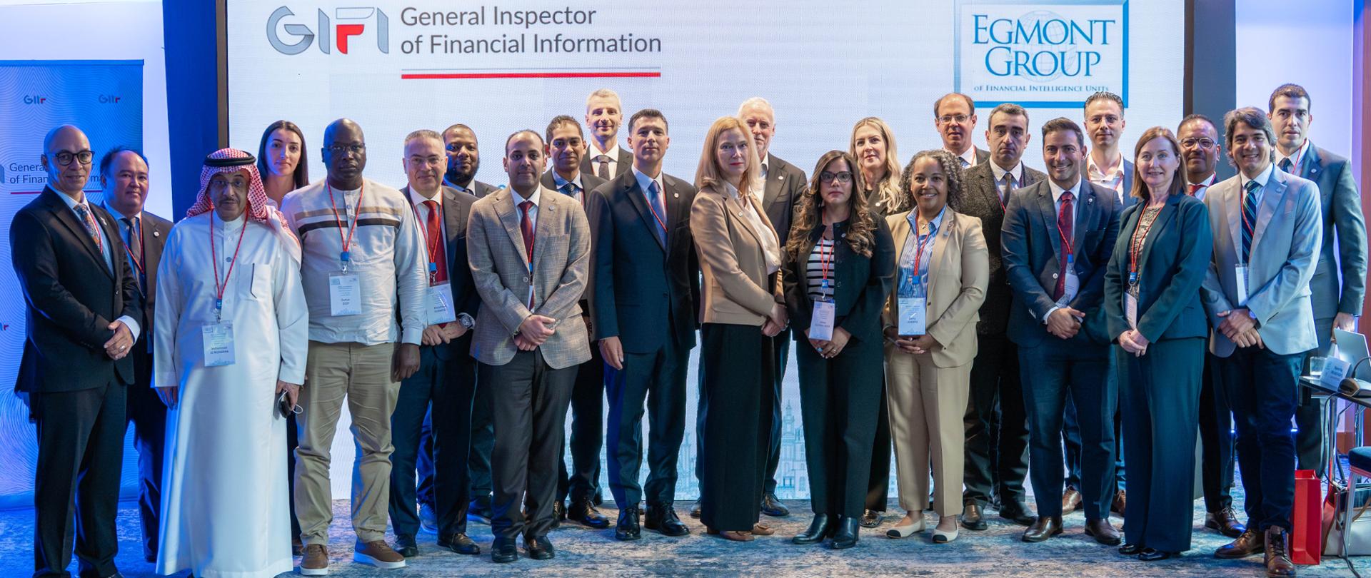 Grupa osób stoi w sali konferencyjnej przed ekranem z napisami „General Inspector of Financial Information” i „Egmont Group”, mając na szyi identyfikatory.