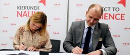 Kobieta w żółtej bluzce w brązowy wzorek i mężczyzna w szarym garniturze siedzą obok siebie przy stole, podpisują dokumenty, za nimi ściana z napisem Kierunek Nauka.