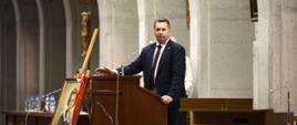 Minister stoi za mównica i mówi do mikrofonu, za nim kamienna ściana, przed nim dużo ludzi.