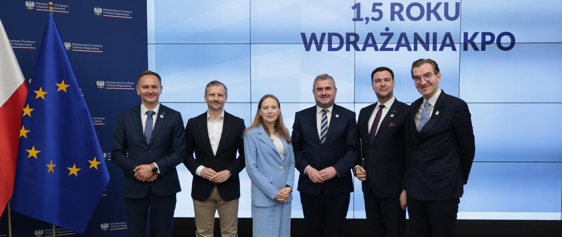 W sali konferencyjnej na scenie stoi obok siebie sześć osób. Trzecia od lewej minister Katarzyna Pełczyńska-Nałęcz, pierwszy od prawej wiceminister Jan Szyszko. Z boku flagi PL i UE. Na ekranie napis: 1,5 roku wdrażania KPO.