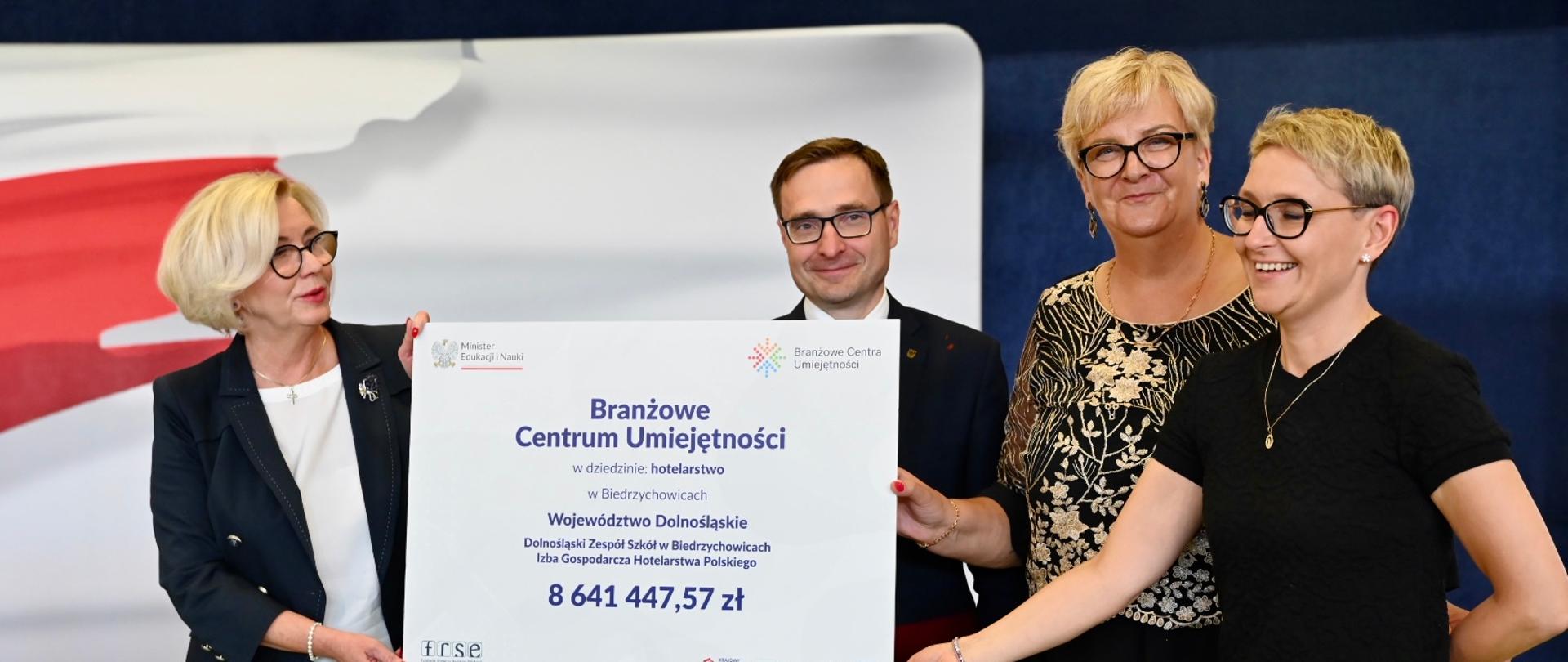Wiceminister Machałek stoi obok mężczyzny w garniturze, kobiety w czarnej sukience i kobiety w czarno-białym ubraniu, wszyscy trzymają wielki symboliczny czek z napisem Branżowe Centrum Umiejętności - 8 641 447,57 zł.